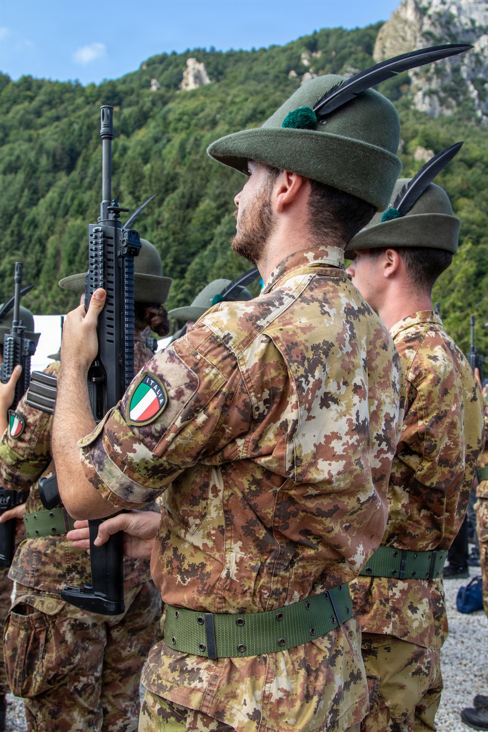 Italian military soldiers, identifiable as Alpini (mountain infantry), are standing in formation during what appears to be a ceremonial parade or military event. The soldiers wear distinctive Alpini felt hats with black feathers and green pompoms, Italian camouflage uniforms (Vegetato pattern), and carry assault rifles. The mountainous, forested backdrop suggests an Alpine or northern Italian setting.