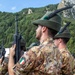 Italian military soldiers, identifiable as Alpini (mountain infantry), are standing in formation during what appears to be a ceremonial parade or military event. The soldiers wear distinctive Alpini felt hats with black feathers and green pompoms, Italian camouflage uniforms (Vegetato pattern), and carry assault rifles. The mountainous, forested backdrop suggests an Alpine or northern Italian setting.
