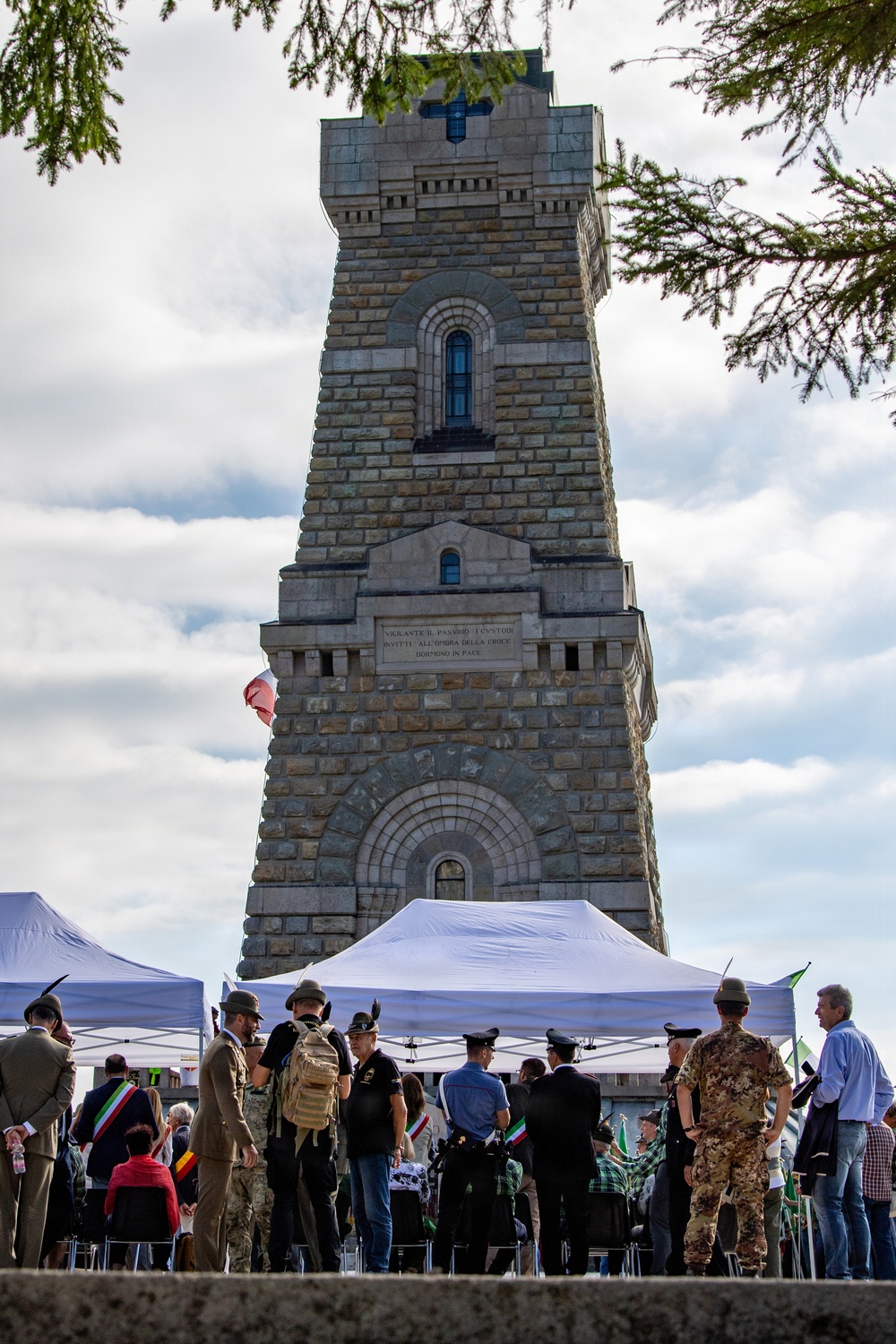 A commemorative ceremony is taking place at the base of a large stone memorial tower, likely an Italian war memorial. The inscription on the tower reads 'VARCANTE IL PASSO I CUSTODI INVITTI ALL'OPERA DELLA CROCE DORMONO IN PACE', and a cross is visible at the top. Military personnel, officials, and civilians are gathered under white canopy tents for what appears to be a formal remembrance event.