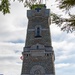 A commemorative ceremony is taking place at the base of a large stone memorial tower, likely an Italian war memorial. The inscription on the tower reads 'VARCANTE IL PASSO I CUSTODI INVITTI ALL'OPERA DELLA CROCE DORMONO IN PACE', and a cross is visible at the top. Military personnel, officials, and civilians are gathered under white canopy tents for what appears to be a formal remembrance event.