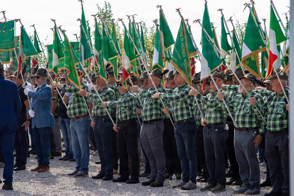 A large formation of uniformed men, likely members of the Italian Alpini (mountain troops veterans association), are standing in rows holding green and Italian tricolor flags at a formal outdoor ceremony. The participants wear matching green-and-black plaid shirts, dark hats with feathers, and carry flags on poles topped with eagle finials. One flag visible reads 'Sezione di Vicenza', indicating this is a regional gathering or national rally of the Alpini association.