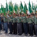 A large formation of uniformed men, likely members of the Italian Alpini (mountain troops veterans association), are standing in rows holding green and Italian tricolor flags at a formal outdoor ceremony. The participants wear matching green-and-black plaid shirts, dark hats with feathers, and carry flags on poles topped with eagle finials. One flag visible reads 'Sezione di Vicenza', indicating this is a regional gathering or national rally of the Alpini association.