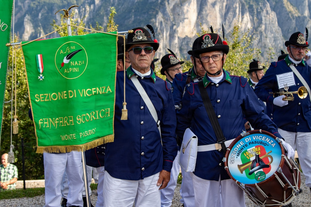 A ceremonial parade or gathering of the Associazione Nazionale Alpini, specifically the 'Fanfara Storica Monte Pasubio' band from the Sezione di Vicenza. Members are dressed in traditional Alpini uniforms consisting of blue jackets, white trousers, and characteristic black hats with feathers. The event appears to take place outdoors against a mountainous backdrop.
