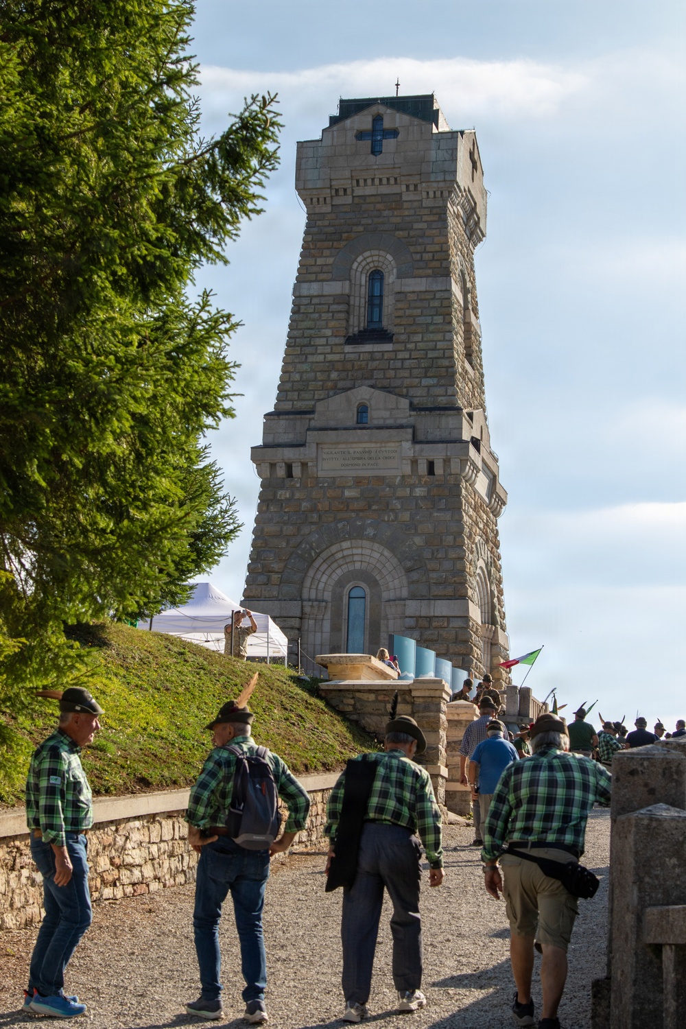 A group of people wearing matching green-and-black plaid shirts and traditional Alpine hats with feathers gather at the base of a tall stone memorial tower featuring a cross near its top. The tower appears to be a war memorial or ossuary, constructed from cut stone blocks with Romanesque-style arched windows and an inscribed plaque. The scene suggests an organized commemorative gathering or veterans' association event.