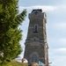 A group of people wearing matching green-and-black plaid shirts and traditional Alpine hats with feathers gather at the base of a tall stone memorial tower featuring a cross near its top. The tower appears to be a war memorial or ossuary, constructed from cut stone blocks with Romanesque-style arched windows and an inscribed plaque. The scene suggests an organized commemorative gathering or veterans' association event.