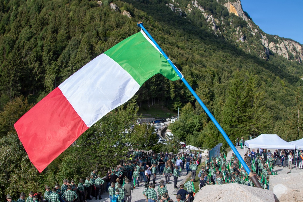 An outdoor gathering in a mountainous Italian setting featuring a large Italian tricolor flag prominently displayed on a blue pole in the foreground. A large crowd of participants, most wearing matching green plaid/checkered shirts and dark hats, are assembled on a paved area surrounded by forested Alpine mountains. The event appears to be an organized ceremonial or commemorative gathering, possibly associated with a regional or national Italian organization.