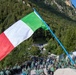 An outdoor gathering in a mountainous Italian setting featuring a large Italian tricolor flag prominently displayed on a blue pole in the foreground. A large crowd of participants, most wearing matching green plaid/checkered shirts and dark hats, are assembled on a paved area surrounded by forested Alpine mountains. The event appears to be an organized ceremonial or commemorative gathering, possibly associated with a regional or national Italian organization.