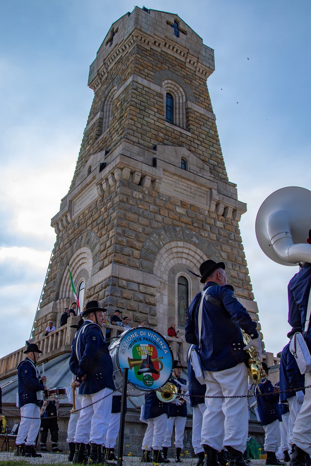 A military or historical band named 'Fanfara Storica - Sezione di Vicenza Monte Pasubio' is performing in front of a large stone memorial tower. The tower appears to be a WWI ossuary or war memorial, likely the Sacrario Militare del Pasubio in northern Italy. Band members are dressed in traditional Alpine-style uniforms with black hats, blue jackets, and white trousers.