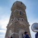A military or historical band named 'Fanfara Storica - Sezione di Vicenza Monte Pasubio' is performing in front of a large stone memorial tower. The tower appears to be a WWI ossuary or war memorial, likely the Sacrario Militare del Pasubio in northern Italy. Band members are dressed in traditional Alpine-style uniforms with black hats, blue jackets, and white trousers.