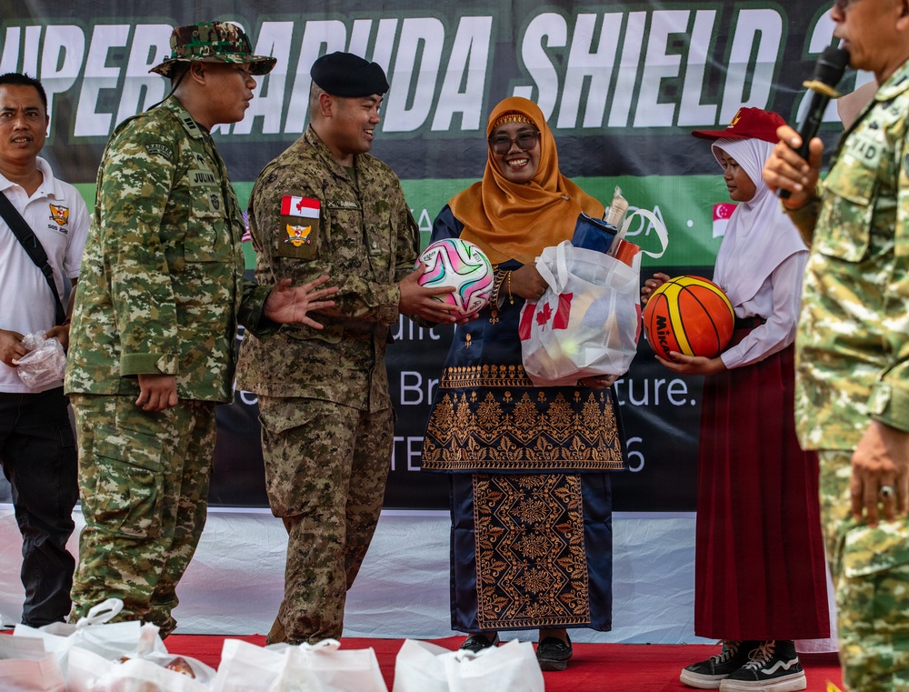 A ceremony at the 'Super Garuda Shield' military exercise where personnel in camouflage uniforms are presenting sports balls and gift bags to civilians, including a woman in traditional Indonesian dress and hijab and a young student. The event appears to be a community outreach or goodwill activity associated with the multinational military exercise. A Canadian flag patch is visible on one soldier's uniform, indicating international participation.