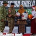 A ceremony at the 'Super Garuda Shield' military exercise where personnel in camouflage uniforms are presenting sports balls and gift bags to civilians, including a woman in traditional Indonesian dress and hijab and a young student. The event appears to be a community outreach or goodwill activity associated with the multinational military exercise. A Canadian flag patch is visible on one soldier's uniform, indicating international participation.