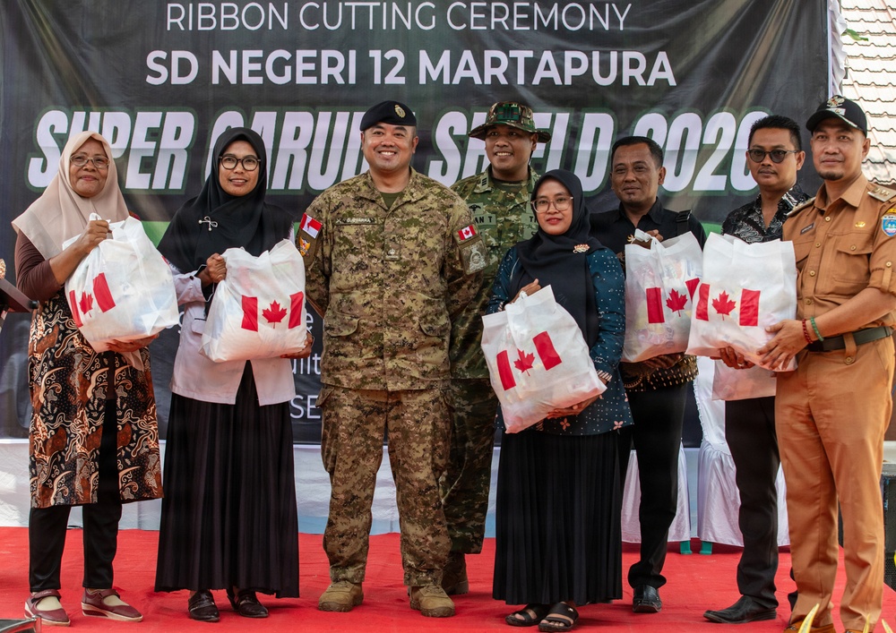 A ribbon cutting ceremony is taking place at SD Negeri 12 Martapura as part of 'Super Garuda Shield 2023,' a military exercise. A group of approximately eight people, including two military personnel in camouflage uniforms and several civilians, are posing together holding white bags bearing the Canadian flag. The event appears to represent a community engagement or humanitarian assistance activity involving Canadian and Indonesian military cooperation.