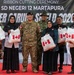 A ribbon cutting ceremony is taking place at SD Negeri 12 Martapura as part of 'Super Garuda Shield 2023,' a military exercise. A group of approximately eight people, including two military personnel in camouflage uniforms and several civilians, are posing together holding white bags bearing the Canadian flag. The event appears to represent a community engagement or humanitarian assistance activity involving Canadian and Indonesian military cooperation.
