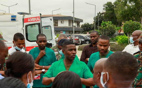 A group of medical and military personnel are gathered in an outdoor briefing or debriefing session near an ambulance. The group includes individuals wearing green surgical scrubs, white lab coats, military camouflage uniforms, and some wearing face masks. The setting appears to be outside a medical facility, likely in a Portuguese-speaking African country based on the partial 'AMBULÂNCIA' text visible on the vehicle.