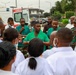 A group of medical and military personnel are gathered in an outdoor briefing or debriefing session near an ambulance. The group includes individuals wearing green surgical scrubs, white lab coats, military camouflage uniforms, and some wearing face masks. The setting appears to be outside a medical facility, likely in a Portuguese-speaking African country based on the partial 'AMBULÂNCIA' text visible on the vehicle.