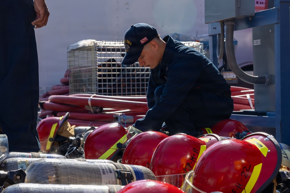 A Navy sailor wearing a cap with 'IWO JIMA' text and a US flag patch is inspecting or organizing firefighting equipment on what appears to be a naval vessel. Multiple red firefighting helmets with yellow reflective strips and SCBA (Self-Contained Breathing Apparatus) air cylinders are laid out in front of him. Red fire hoses and a wire cage are visible in the background.