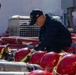 A Navy sailor wearing a cap with 'IWO JIMA' text and a US flag patch is inspecting or organizing firefighting equipment on what appears to be a naval vessel. Multiple red firefighting helmets with yellow reflective strips and SCBA (Self-Contained Breathing Apparatus) air cylinders are laid out in front of him. Red fire hoses and a wire cage are visible in the background.
