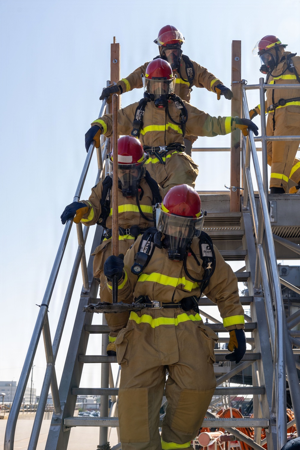 A group of firefighters in full turnout gear and SCBA (Self-Contained Breathing Apparatus) equipment are climbing a metal training staircase structure. The personnel are wearing tan/gold bunker gear with high-visibility yellow reflective stripes and red helmets. This appears to be a firefighter training exercise conducted at an outdoor industrial or training facility.