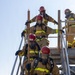 A group of firefighters in full turnout gear and SCBA (Self-Contained Breathing Apparatus) equipment are climbing a metal training staircase structure. The personnel are wearing tan/gold bunker gear with high-visibility yellow reflective stripes and red helmets. This appears to be a firefighter training exercise conducted at an outdoor industrial or training facility.