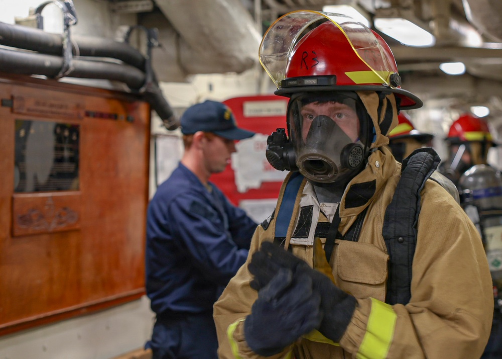 A person in full firefighting gear, including a red helmet marked 'R3', a full-face respirator mask, tan turnout gear with reflective yellow stripes, and black gloves, stands in what appears to be the interior of a naval vessel. In the background, another individual in a navy blue uniform and cap is visible near a wooden panel with mounted equipment. Additional personnel in red helmets are partially visible in the far background.