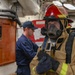 A person in full firefighting gear, including a red helmet marked 'R3', a full-face respirator mask, tan turnout gear with reflective yellow stripes, and black gloves, stands in what appears to be the interior of a naval vessel. In the background, another individual in a navy blue uniform and cap is visible near a wooden panel with mounted equipment. Additional personnel in red helmets are partially visible in the far background.