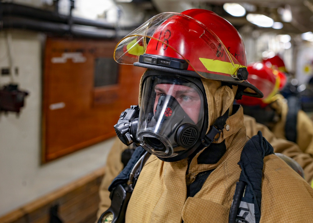 A person wearing full firefighting gear, including a red helmet marked with the number '13', a full-face respirator/SCBA mask, and a tan/gold fire-resistant suit, is visible in the foreground. The setting appears to be the interior of a naval vessel or industrial facility, with additional similarly-equipped personnel visible in the background. The image appears to document a firefighting drill or emergency response exercise.