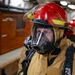 A person wearing full firefighting gear, including a red helmet marked with the number '13', a full-face respirator/SCBA mask, and a tan/gold fire-resistant suit, is visible in the foreground. The setting appears to be the interior of a naval vessel or industrial facility, with additional similarly-equipped personnel visible in the background. The image appears to document a firefighting drill or emergency response exercise.
