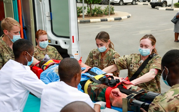 A medical evacuation scenario or training exercise is taking place outside an ambulance, where military personnel in camouflage uniforms and civilian medical staff in white lab coats are jointly transferring a patient on a stretcher. Multiple personnel wearing surgical masks are collaborating on the patient handoff, suggesting a joint military-civilian medical cooperation exercise. The setting appears to be a hospital or medical facility entrance in what appears to be an African country.