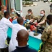 A medical evacuation scenario or training exercise is taking place outside an ambulance, where military personnel in camouflage uniforms and civilian medical staff in white lab coats are jointly transferring a patient on a stretcher. Multiple personnel wearing surgical masks are collaborating on the patient handoff, suggesting a joint military-civilian medical cooperation exercise. The setting appears to be a hospital or medical facility entrance in what appears to be an African country.