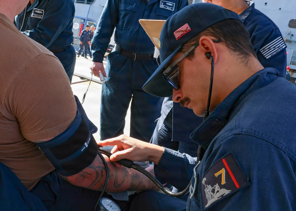 A Navy corpsman or medical personnel in dark blue military uniform is taking a blood pressure reading on a patient's arm using a sphygmomanometer and stethoscope. The setting appears to be an outdoor naval facility or pier, with multiple other personnel in similar dark blue uniforms visible in the background. The medical provider is wearing a cap with an American flag patch and a unit patch with the number '7' on their sleeve.