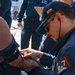 A Navy corpsman or medical personnel in dark blue military uniform is taking a blood pressure reading on a patient's arm using a sphygmomanometer and stethoscope. The setting appears to be an outdoor naval facility or pier, with multiple other personnel in similar dark blue uniforms visible in the background. The medical provider is wearing a cap with an American flag patch and a unit patch with the number '7' on their sleeve.