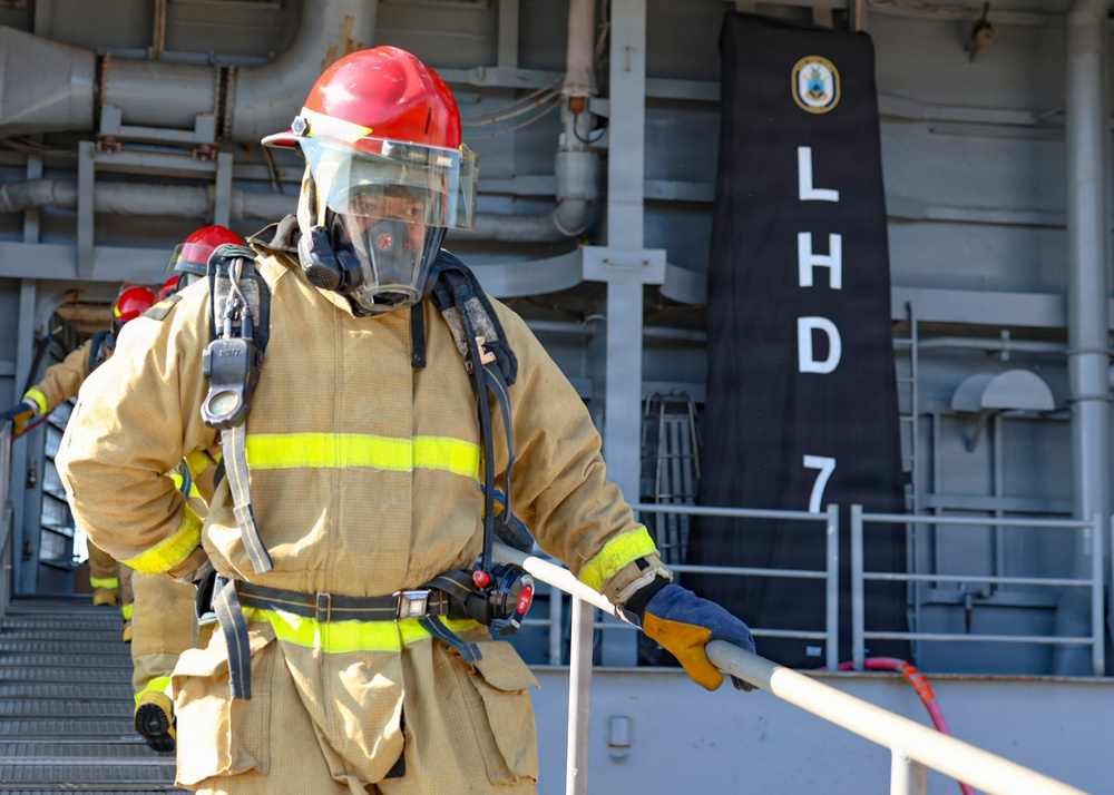 The image shows naval firefighters in full firefighting gear aboard a U.S. Navy amphibious assault ship designated LHD-7, identified as USS Iwo Jima. Personnel are wearing full turnout gear including SCBA (Self-Contained Breathing Apparatus), red helmets, face masks, and high-visibility reflective jackets. This appears to be a firefighting drill or training exercise conducted on the ship's deck.