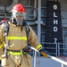 The image shows naval firefighters in full firefighting gear aboard a U.S. Navy amphibious assault ship designated LHD-7, identified as USS Iwo Jima. Personnel are wearing full turnout gear including SCBA (Self-Contained Breathing Apparatus), red helmets, face masks, and high-visibility reflective jackets. This appears to be a firefighting drill or training exercise conducted on the ship's deck.