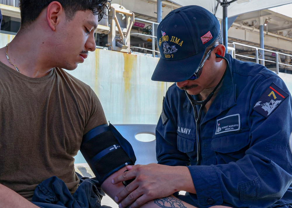 A U.S. Navy medical personnel identified as Robert Maruffo is taking a blood pressure reading on a patient aboard a naval vessel. The medic is wearing a USS Iwo Jima (LHD-7) cap and U.S. Navy uniform with a stethoscope. The scene appears to be an outdoor deck area of the ship.