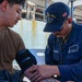 A U.S. Navy medical personnel identified as Robert Maruffo is taking a blood pressure reading on a patient aboard a naval vessel. The medic is wearing a USS Iwo Jima (LHD-7) cap and U.S. Navy uniform with a stethoscope. The scene appears to be an outdoor deck area of the ship.