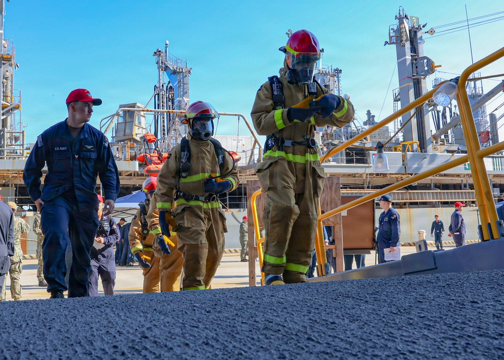 The image depicts a military firefighting drill or training exercise at what appears to be a naval industrial facility or shipyard. Several personnel in full firefighting gear — including turnout suits, SCBA masks, and red helmets — are ascending a ramp alongside U.S. Navy personnel in blue uniforms. The background shows large industrial structures, yellow safety railings, and additional observers and participants.