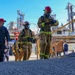 The image depicts a military firefighting drill or training exercise at what appears to be a naval industrial facility or shipyard. Several personnel in full firefighting gear — including turnout suits, SCBA masks, and red helmets — are ascending a ramp alongside U.S. Navy personnel in blue uniforms. The background shows large industrial structures, yellow safety railings, and additional observers and participants.