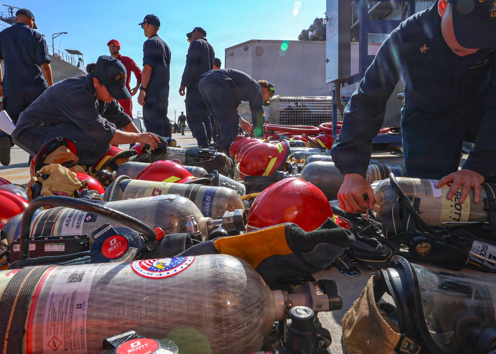 Military or naval personnel in dark blue uniforms are handling and organizing a large collection of Scott Aviation SCBA (Self-Contained Breathing Apparatus) tanks and red firefighting helmets on an outdoor surface. The equipment appears to be part of a firefighting or damage control drill, likely aboard or near a naval vessel. Multiple personnel are actively inspecting, tagging, or sorting the breathing apparatus cylinders.