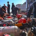 Military or naval personnel in dark blue uniforms are handling and organizing a large collection of Scott Aviation SCBA (Self-Contained Breathing Apparatus) tanks and red firefighting helmets on an outdoor surface. The equipment appears to be part of a firefighting or damage control drill, likely aboard or near a naval vessel. Multiple personnel are actively inspecting, tagging, or sorting the breathing apparatus cylinders.