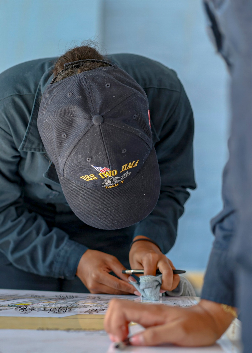 A person wearing a dark navy baseball cap embroidered with 'USS IWO JIMA LHD 7' is bent over a table, writing or marking on a document or map. The individual is dressed in dark military-style clothing and appears to be engaged in a task involving paperwork. A second person is partially visible in the foreground right.