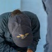 A person wearing a dark navy baseball cap embroidered with 'USS IWO JIMA LHD 7' is bent over a table, writing or marking on a document or map. The individual is dressed in dark military-style clothing and appears to be engaged in a task involving paperwork. A second person is partially visible in the foreground right.