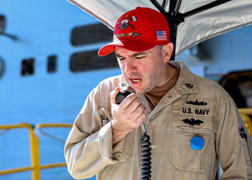 A U.S. Navy servicemember in tan/khaki uniform is speaking into a handheld microphone, likely making an announcement or address. He is wearing a red baseball cap with 'USS IWO JIMA' text and an American flag patch, along with submarine warfare qualification badges on his uniform. The setting appears to be dockside or aboard a naval vessel.