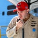 A U.S. Navy servicemember in tan/khaki uniform is speaking into a handheld microphone, likely making an announcement or address. He is wearing a red baseball cap with 'USS IWO JIMA' text and an American flag patch, along with submarine warfare qualification badges on his uniform. The setting appears to be dockside or aboard a naval vessel.