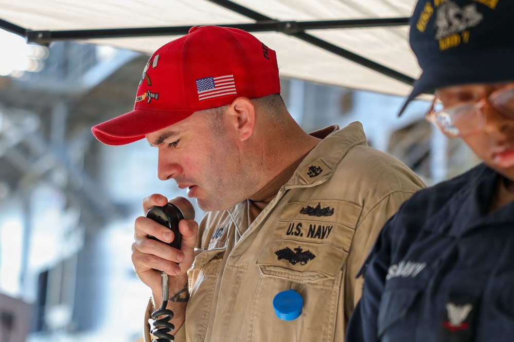 A U.S. Navy servicemember in khaki uniform is speaking into a corded microphone, likely making an announcement or address. He is wearing a red baseball cap with an American flag patch and Navy insignia. A second person in a dark Navy uniform and blue cap is partially visible on the right side of the frame.