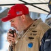 A U.S. Navy servicemember in khaki uniform is speaking into a corded microphone, likely making an announcement or address. He is wearing a red baseball cap with an American flag patch and Navy insignia. A second person in a dark Navy uniform and blue cap is partially visible on the right side of the frame.