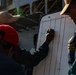 Three individuals are gathered around a whiteboard mounted outdoors, with one person actively writing on it using a marker. The board appears to be a tracking or accountability chart with columns labeled 'IN', 'OUT', and 'LOC'. The setting appears to be near a large naval or industrial vessel in the background.