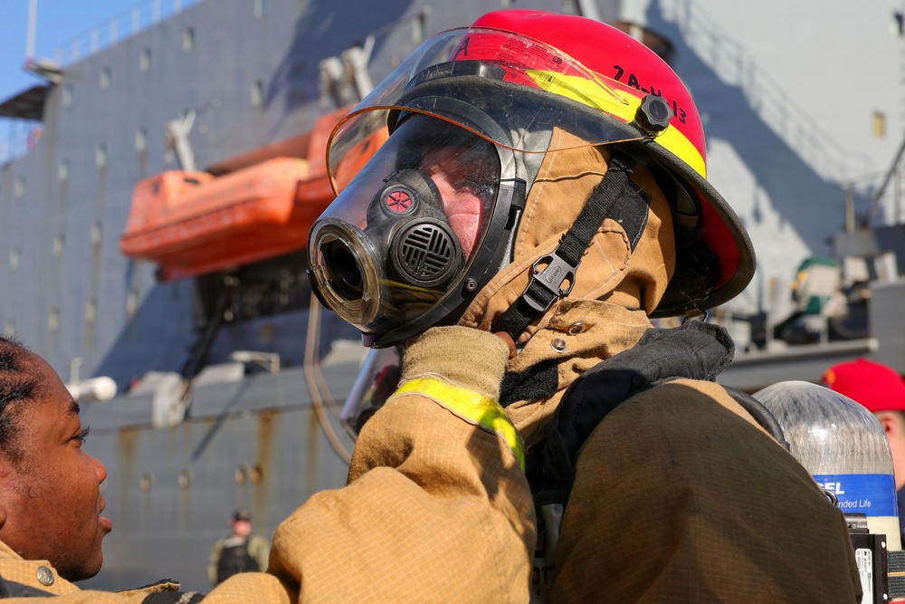 The image shows personnel in firefighting gear, likely aboard or near a naval vessel. One individual wearing a red and yellow firefighting helmet with a full-face respirator mask is being assisted by another person in adjusting or securing the equipment. A large gray naval ship with an orange lifeboat is visible in the background.