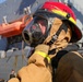 The image shows personnel in firefighting gear, likely aboard or near a naval vessel. One individual wearing a red and yellow firefighting helmet with a full-face respirator mask is being assisted by another person in adjusting or securing the equipment. A large gray naval ship with an orange lifeboat is visible in the background.