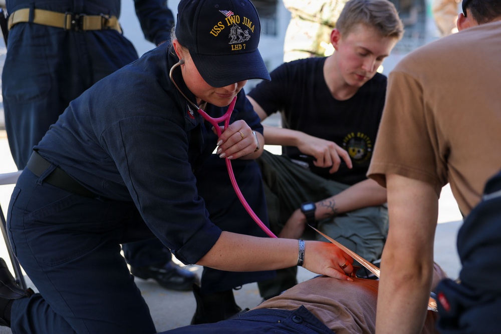 A U.S. Navy medical personnel wearing a USS Iwo Jima (LHD 7) cap is using a pink stethoscope to assess a simulated casualty during what appears to be a medical training exercise. Multiple personnel are attending to the patient, who has simulated blood (red substance) visible on their torso. The scene takes place outdoors on what appears to be a ship deck or military installation.