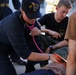 A U.S. Navy medical personnel wearing a USS Iwo Jima (LHD 7) cap is using a pink stethoscope to assess a simulated casualty during what appears to be a medical training exercise. Multiple personnel are attending to the patient, who has simulated blood (red substance) visible on their torso. The scene takes place outdoors on what appears to be a ship deck or military installation.
